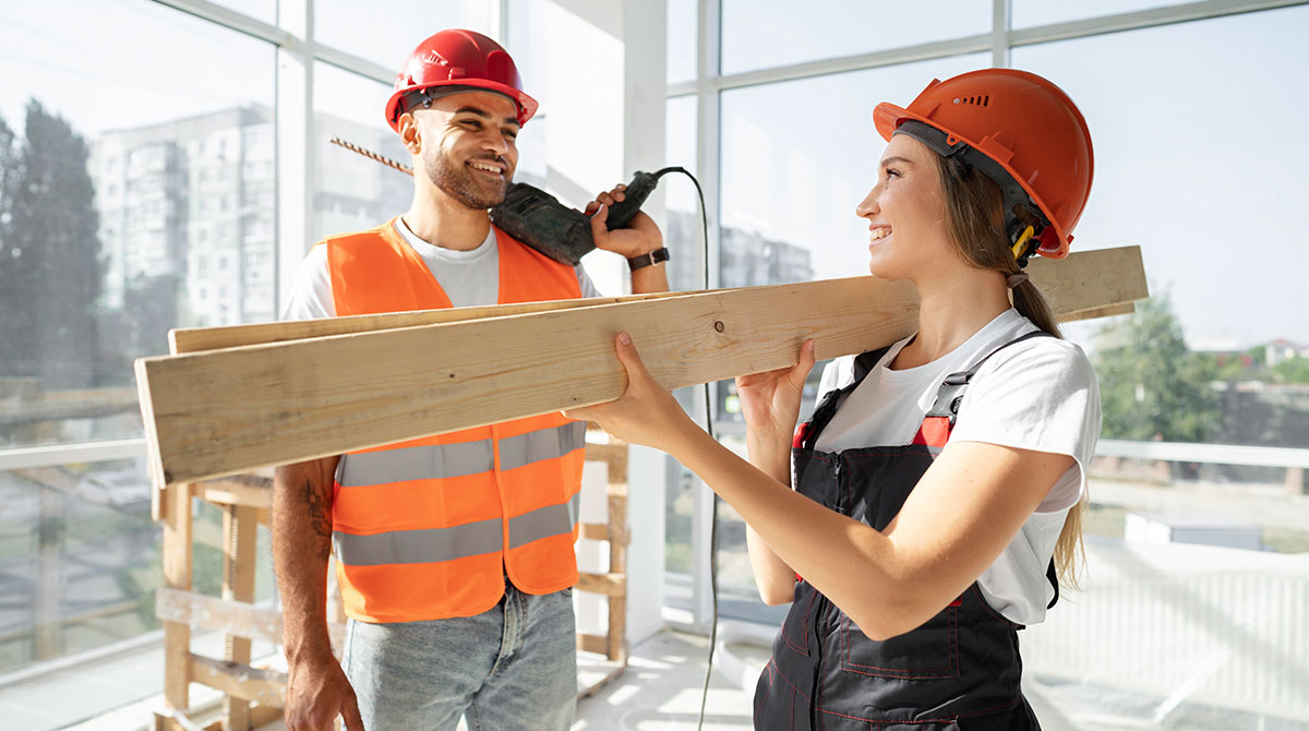 Construction workers smiling and collaborating, one holding a wooden plank and the other with a power tool, in a bright, modern renovation space.